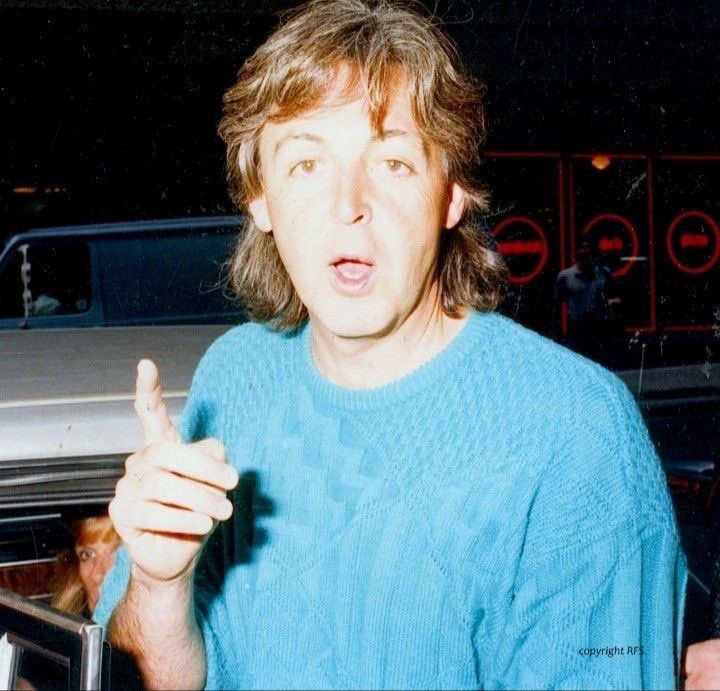 a man in a blue sweater pointing to the left with his finger up while standing next to a car