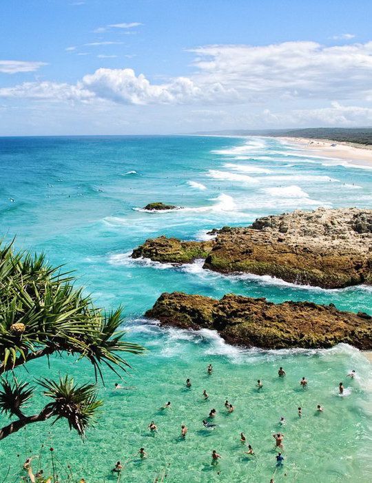 many people are swimming in the ocean near some rocks and trees, while others swim in the water