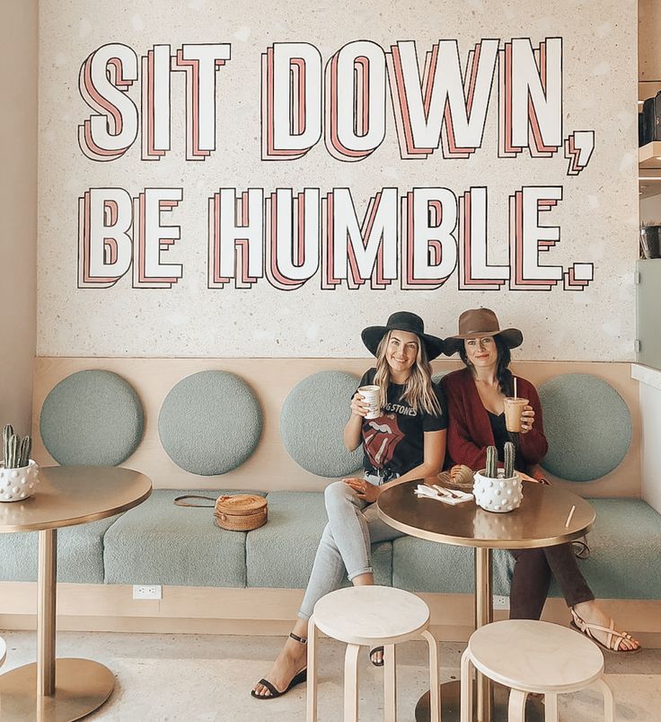 two women sitting at a table in front of a sign that says sit down, be humble