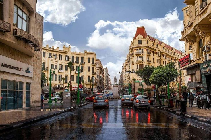 cars driving down a wet street with buildings in the background and people walking on the sidewalk