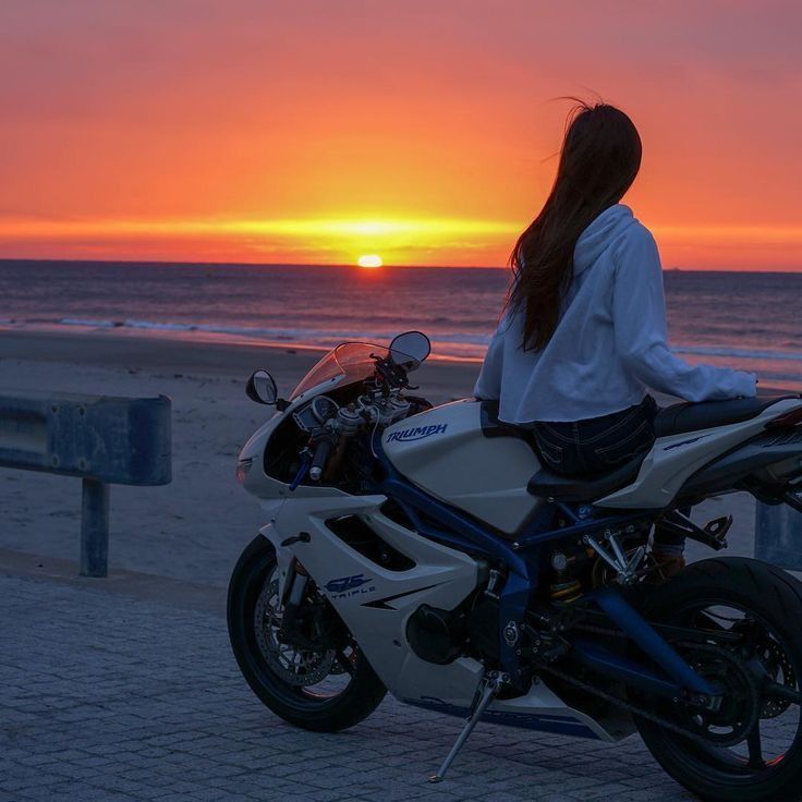 a woman sitting on top of a motorcycle near the ocean at sunset or sunrise time