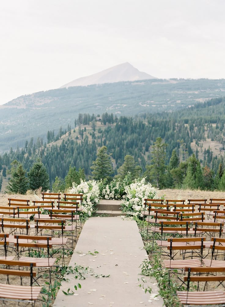 an outdoor ceremony set up with wooden chairs and flowers on the aisle, overlooking mountains