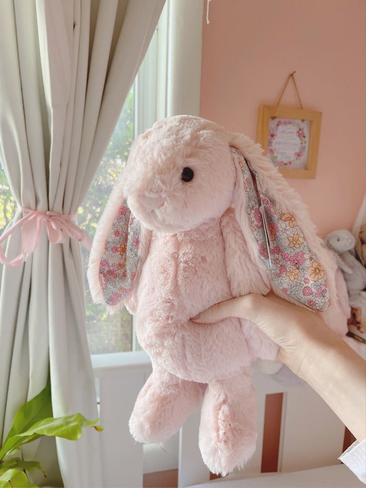 a pink stuffed bunny is held up by someone's hand in front of a window