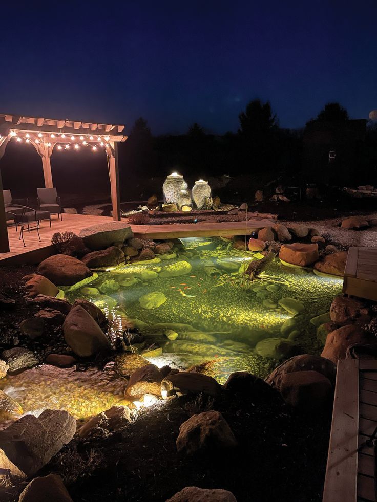 an outdoor pond lit up at night with lights on the water and rocks surrounding it