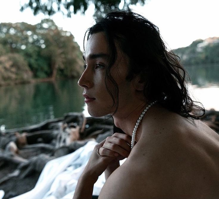 a shirtless man with long hair and pearls on his neck sitting by the water