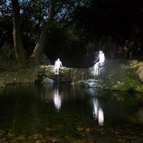 two people standing on rocks in the middle of a stream at night with their lights on