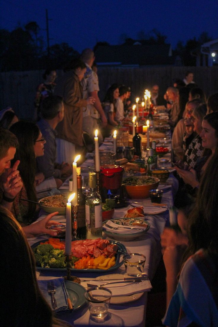 a group of people sitting around a table with food and candles on it at night