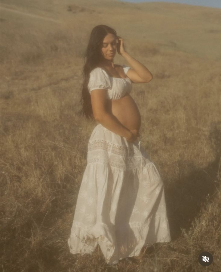 a pregnant woman in a white dress is standing in a field and talking on her cell phone