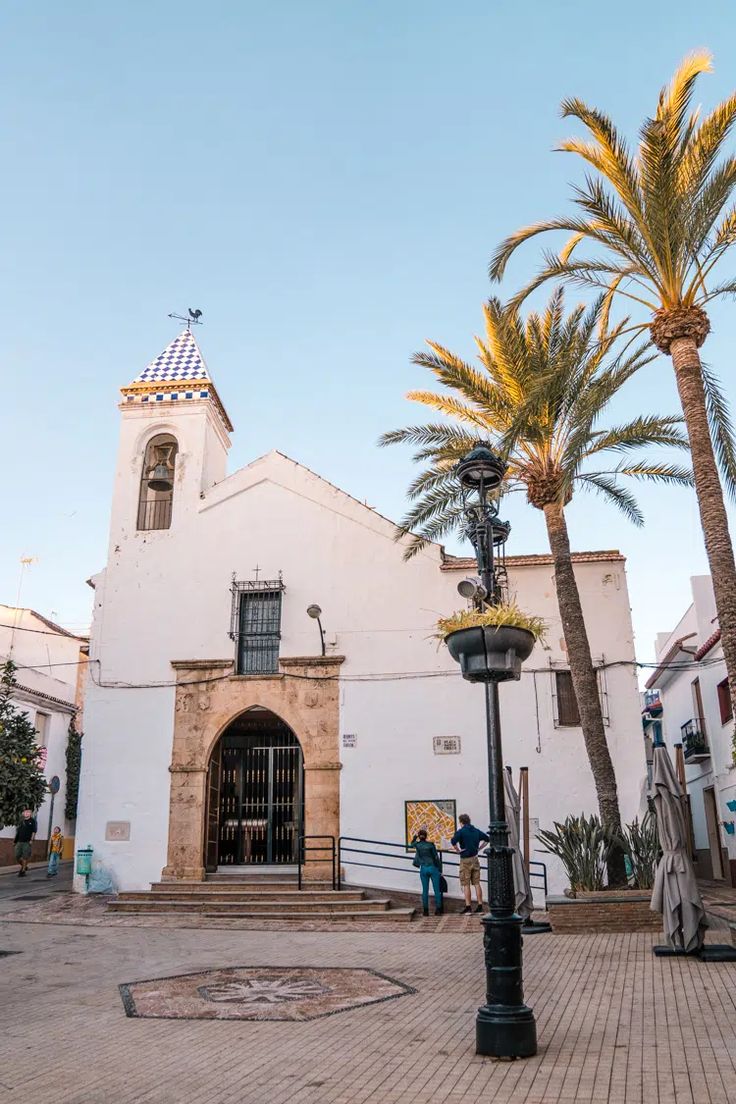 a white church with palm trees in front of it and people walking around the building