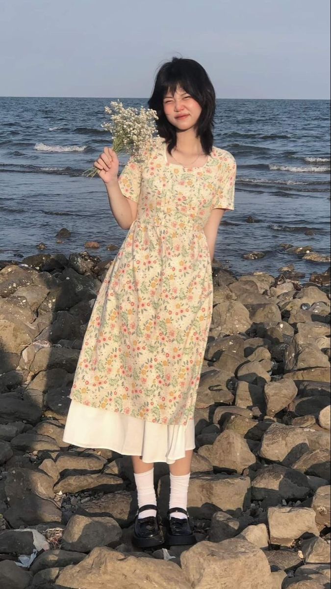 a woman standing on top of a rocky beach next to the ocean holding a bouquet of flowers