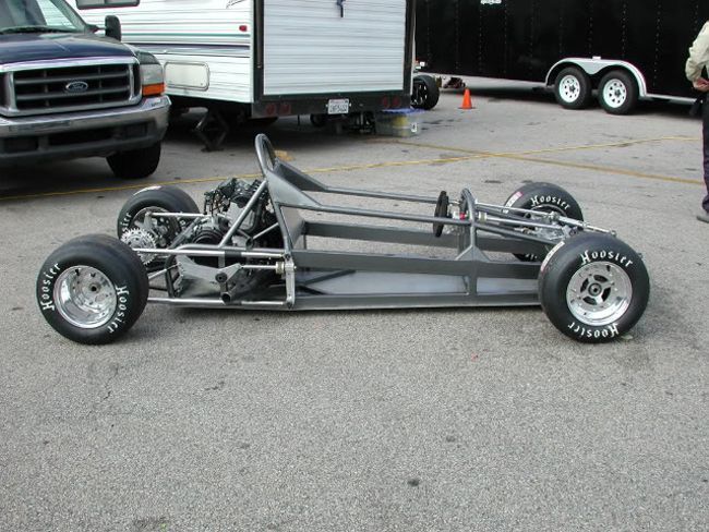 a silver race car sitting in the middle of a parking lot next to a trailer