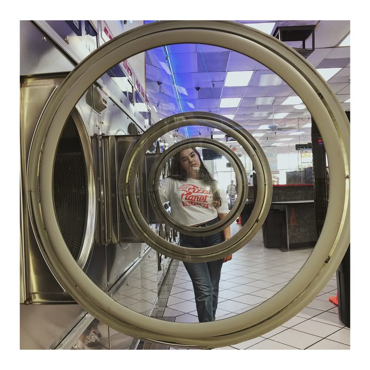 a woman standing in front of a dryer looking at her reflection through the door