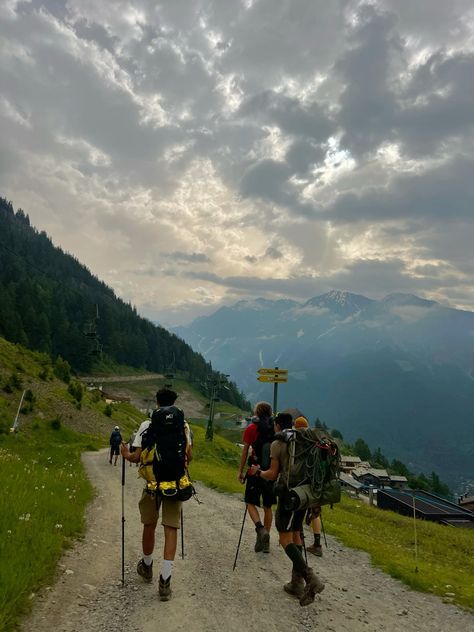 three people with backpacks walking up a dirt road in the mountains on a cloudy day