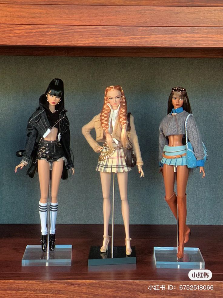 three barbie dolls are standing on display in a wooden case, one is wearing a short skirt and the other has long hair