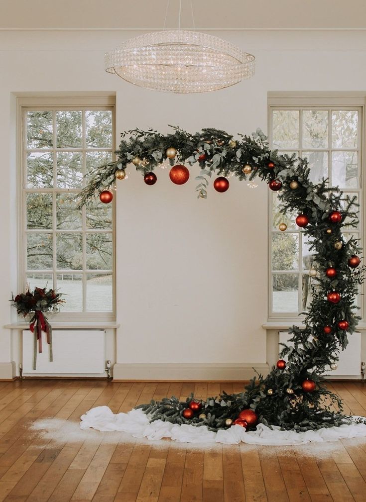 a christmas wreath with ornaments hanging from it on a wooden floor in front of two windows