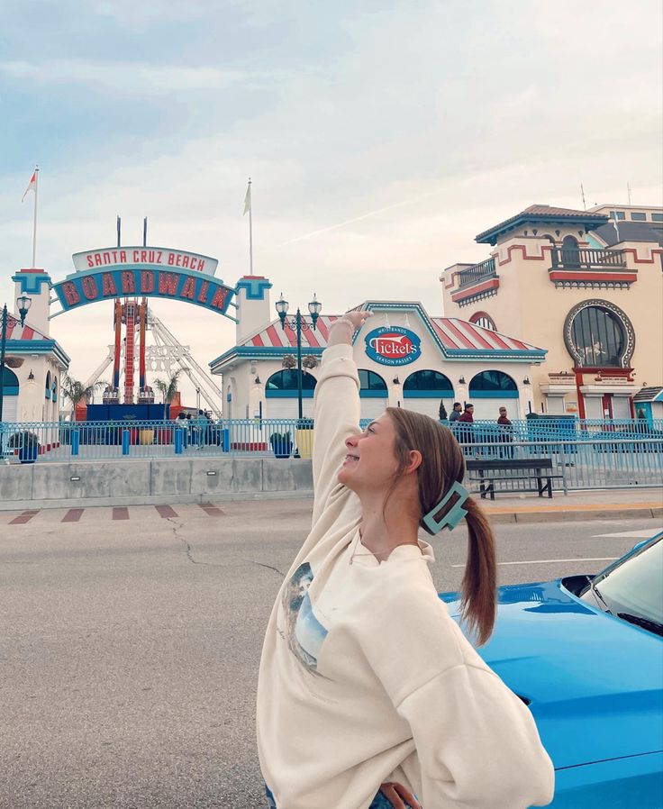 a woman standing next to a blue car in front of a roller coaster at an amusement park