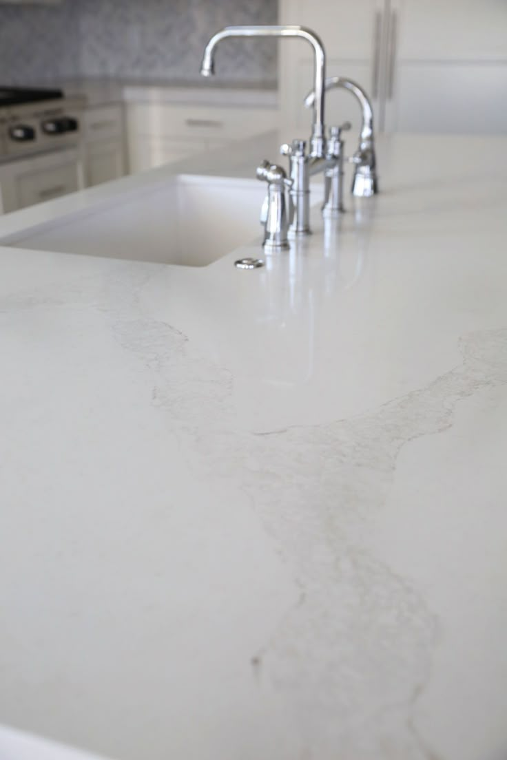 a white kitchen counter top with two faucets on the sink and an oven in the background