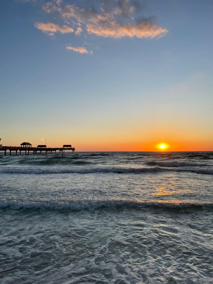 the sun is setting over the ocean with waves crashing in front of it and a pier stretching out into the distance