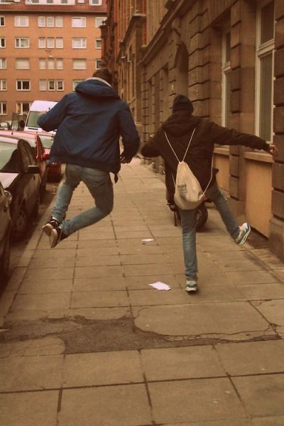 two people are running down the sidewalk in front of parked cars and one person is jumping