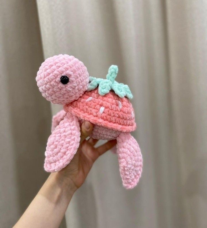 a small pink crocheted stuffed animal with a green leaf on its head is held up in front of a white curtain