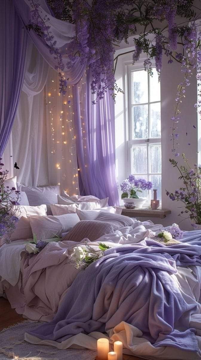 a bed with purple sheets and pillows in front of a window filled with flowers next to two candles