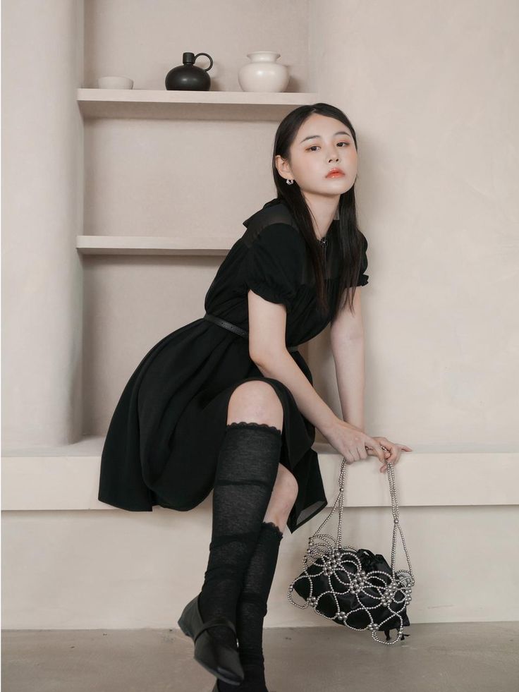 a woman in black dress and knee high socks leaning against wall with handbag on floor