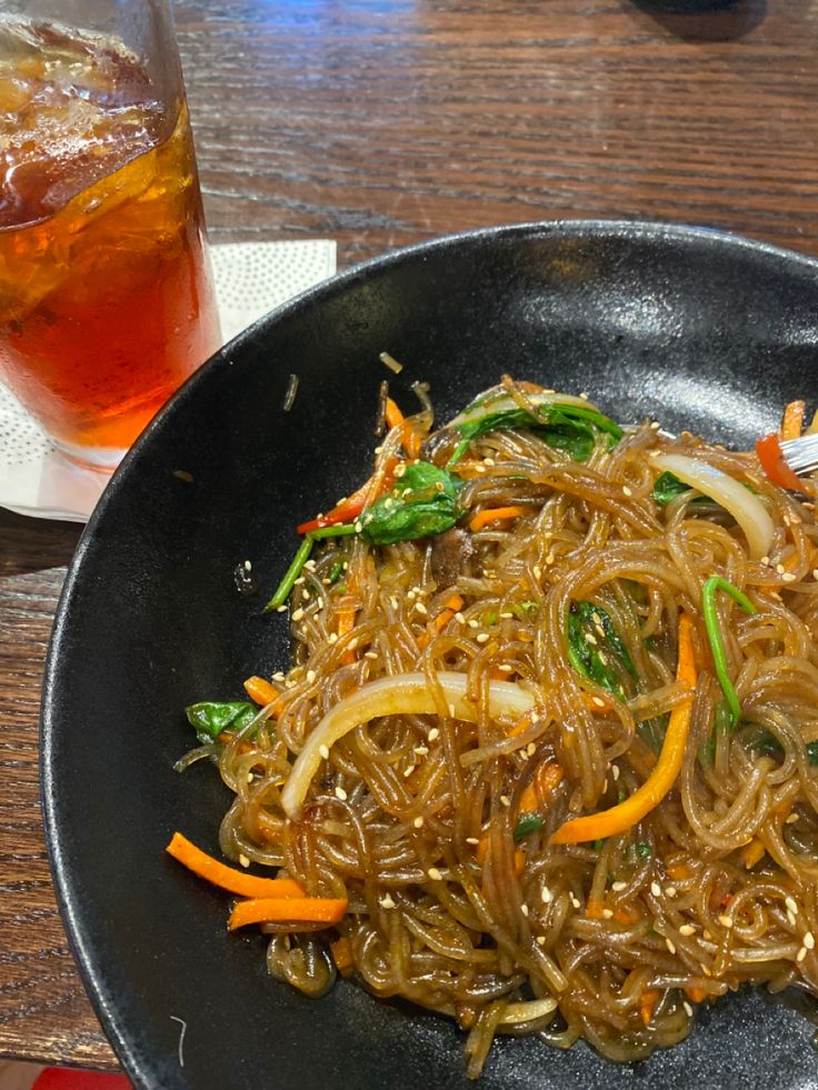 a black plate topped with noodles and veggies next to a glass of soda