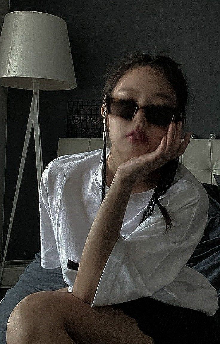 a woman sitting on top of a bed with her hand under her chin and wearing sunglasses