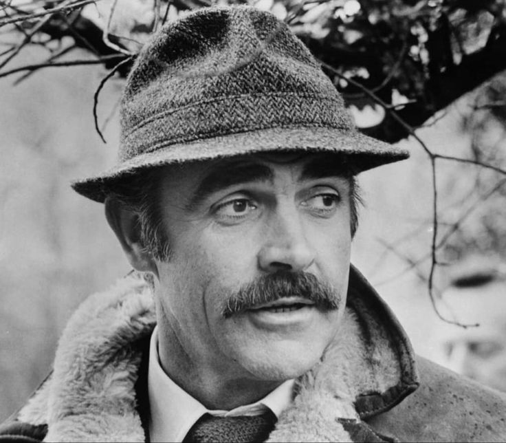 black and white photograph of a man wearing a hat, coat, and tie with a moustache on his face