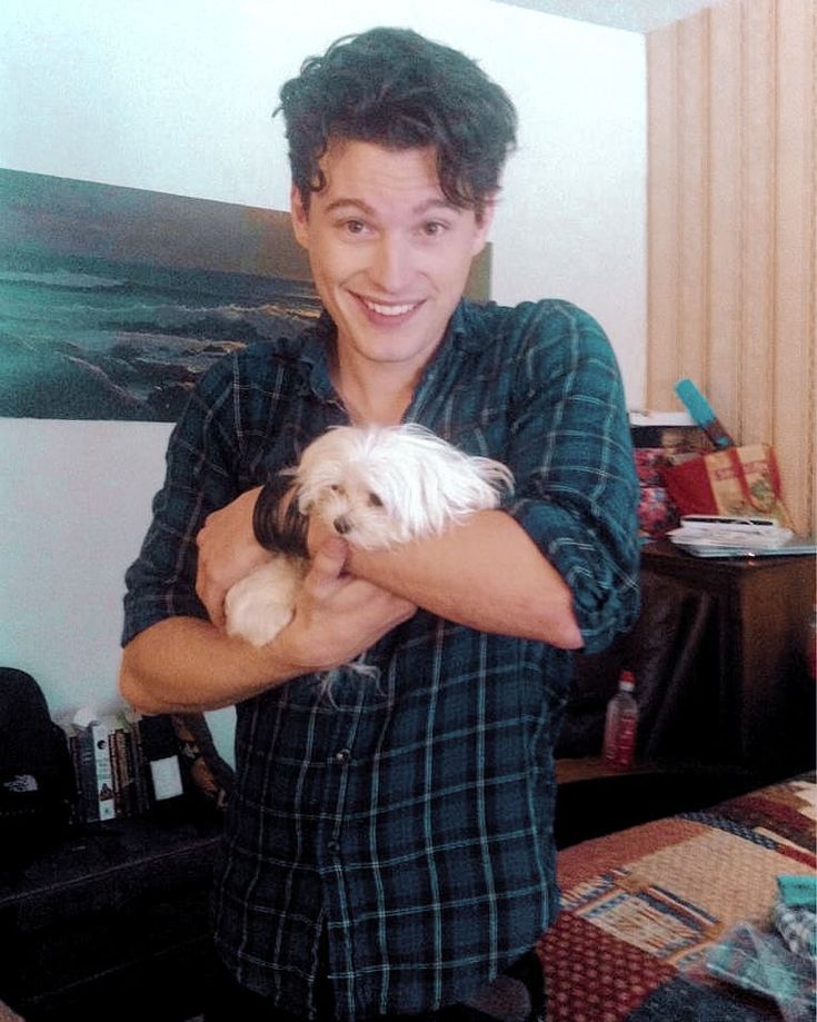 a man holding a small white dog in his arms while standing next to a bed