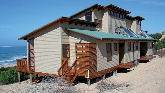 a house on the beach with a green roof and stairs leading up to it's second floor