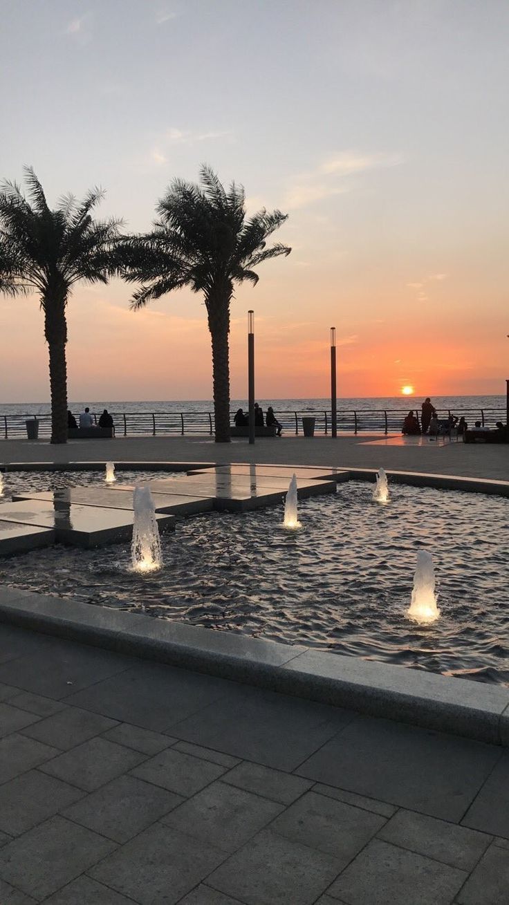 the sun is setting behind two palm trees in front of an outdoor fountain with water cascading