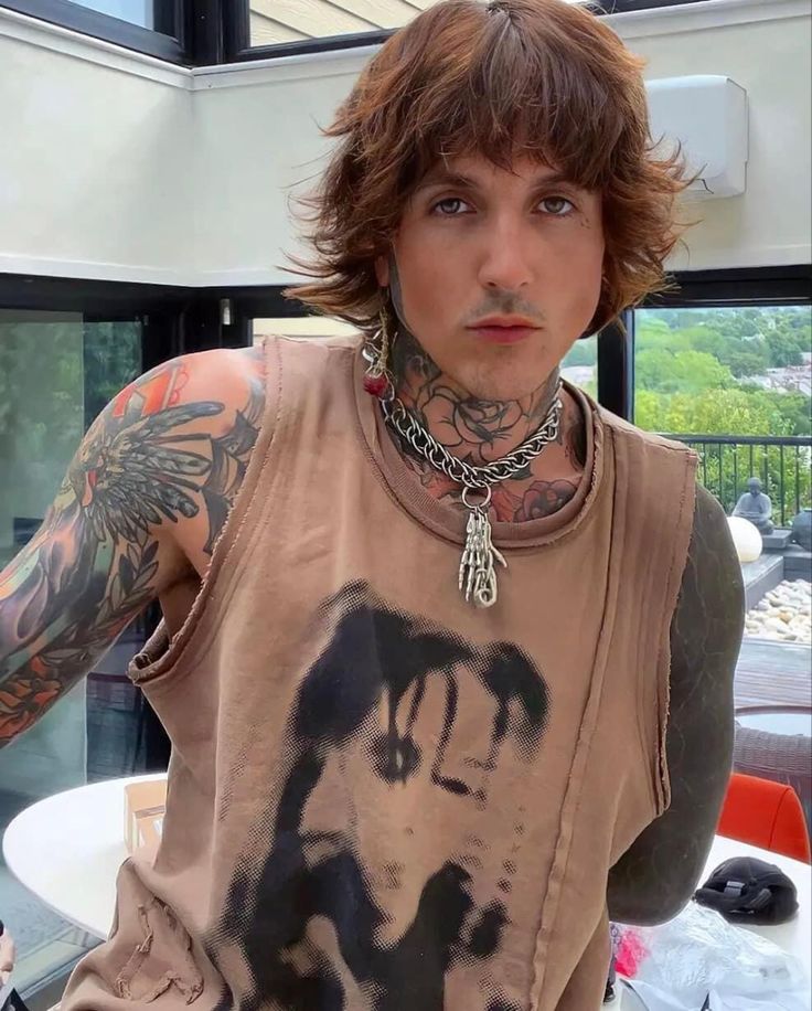 a man with long hair and tattoos standing in front of a window looking at the camera