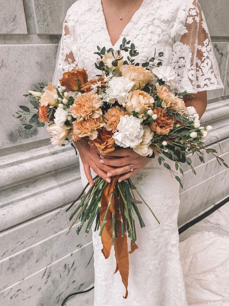 a woman holding a bouquet of flowers in front of a stone building with white and orange colors