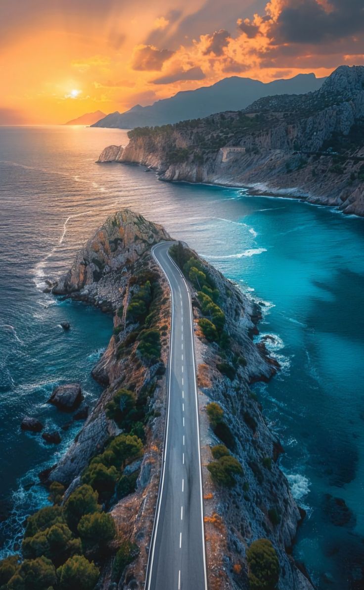 an aerial view of a road near the ocean and mountains at sunset with clouds in the sky