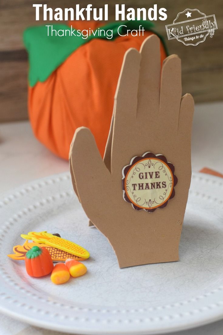 a paper thanksgiving card on a plate with pumpkins in the background and a hand that says give thanks