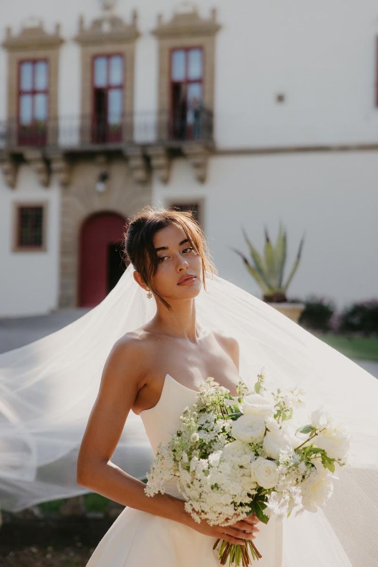 a woman wearing a wedding dress holding a bouquet of flowers in front of a building