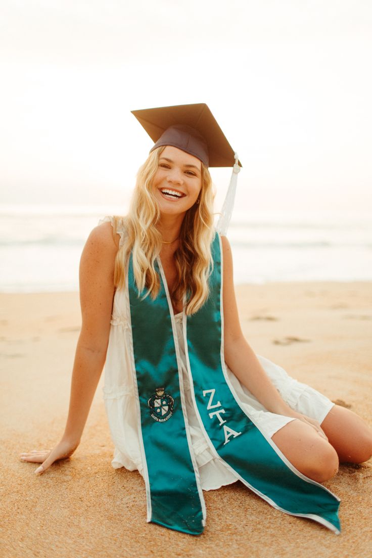 a woman sitting in the sand wearing a graduation cap and gown