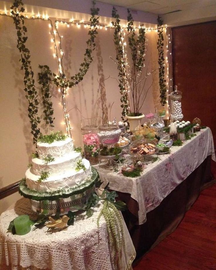 a table topped with lots of cake next to a wall covered in lights and greenery