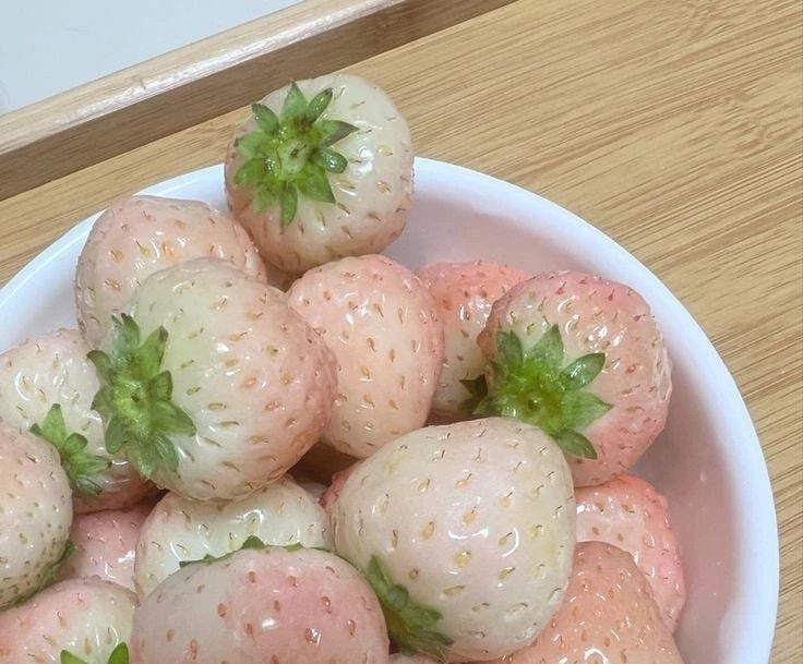 a white bowl filled with lots of strawberries on top of a wooden table next to a window