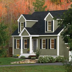 a green house with white trim and black roof in front of some trees that have orange leaves on them