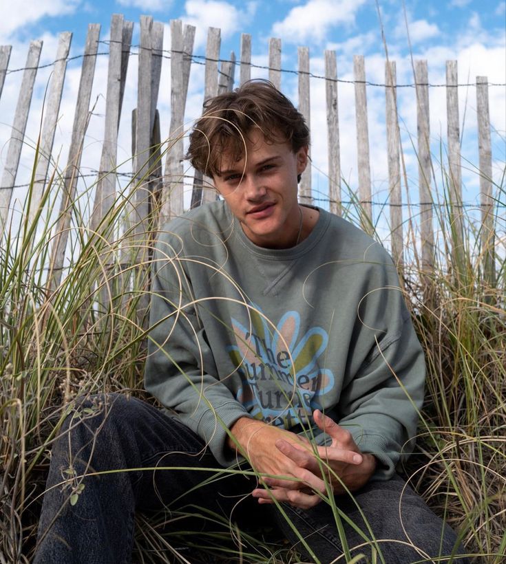 a young man sitting in the sand with his hand on his chest and looking at the camera