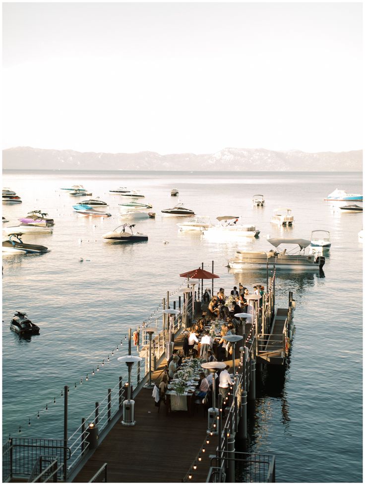 a pier with boats in the water and people sitting at tables on top of it