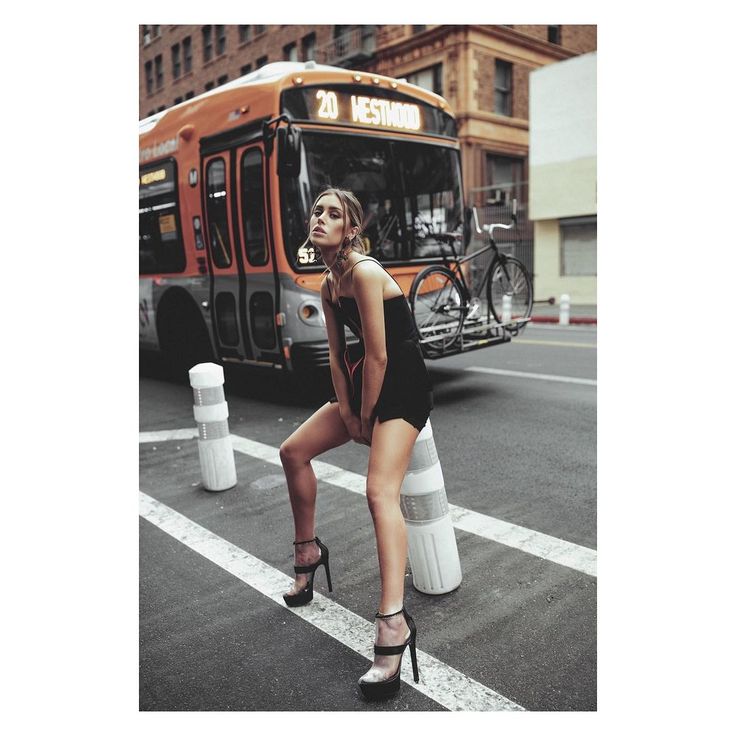 a woman standing on the side of a road in front of a bus with her legs crossed