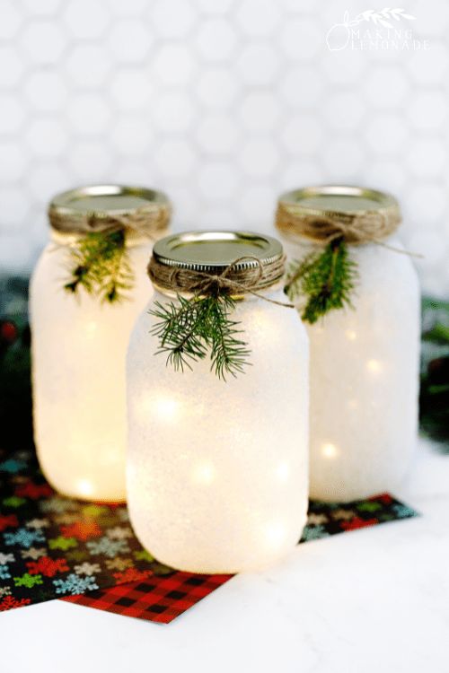 three mason jars filled with white lights on top of a christmas table cloth covered in snowflakes