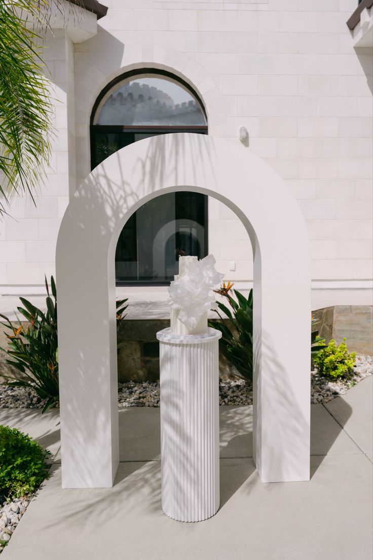 a white vase with flowers in it sitting on the sidewalk next to an arch and palm tree