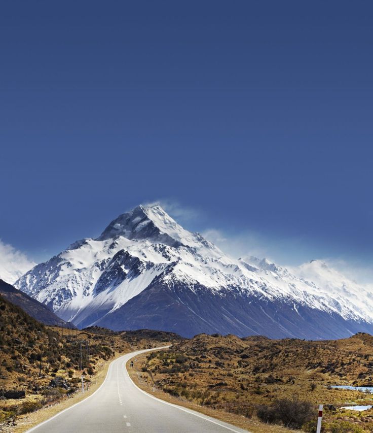 an empty road in the middle of nowhere with a snow covered mountain behind it and blue sky above