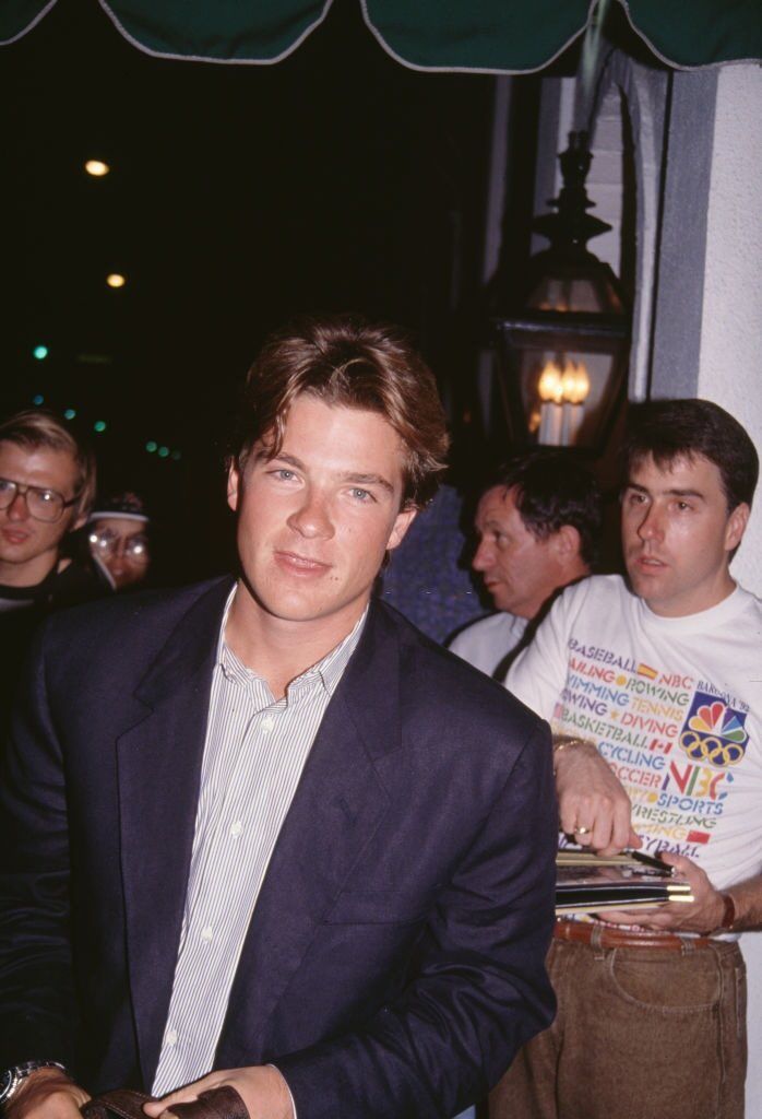 a man in a suit sitting at a table with other people around him and looking into the camera