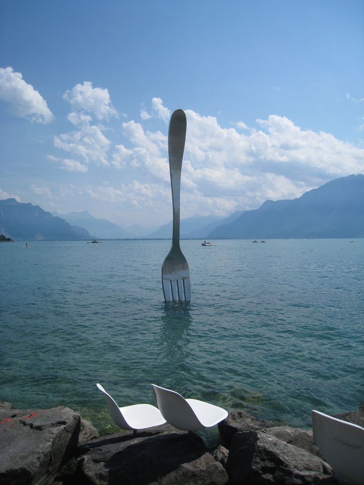 a fork sticking out of the water with two chairs in front of it and mountains in the background