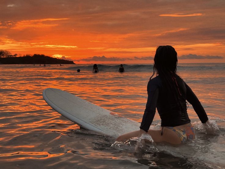 a woman is sitting on her surfboard in the water as the sun goes down